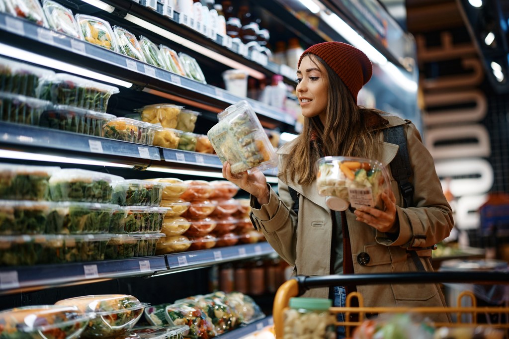 Woman reading a food label in a grocery store aisle, holding two pre-packaged meals