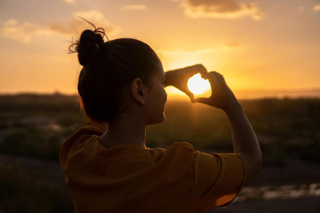 Woman forming a heart shape with her hands around the setting sun, symbolizing gratitude, love, or happiness during golden hour.