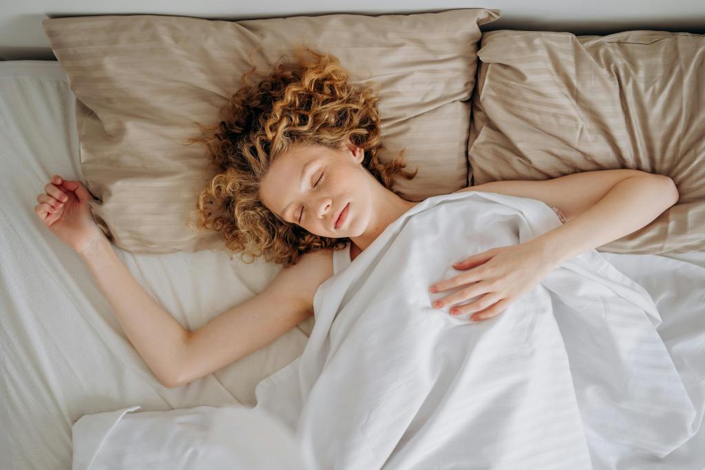 A young woman with curly blonde hair sleeping peacefully in bed, lying on beige pillows and wrapped in a white blanket.